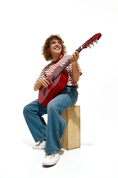 Portrait of happy young man, musician wearing casual clothes, sitting on wooden box and playing guitar isolated on white background. Concept of music, festival, concert, hobby, talent, performance - Fotó, kép