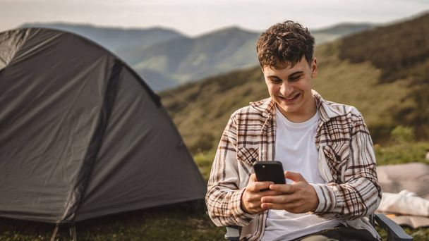 Νεαρό αγόρι κάθεται μπροστά σκηνή χρησιμοποιούν το κινητό τηλέφωνο στο βουνό - Φωτογραφία, εικόνα