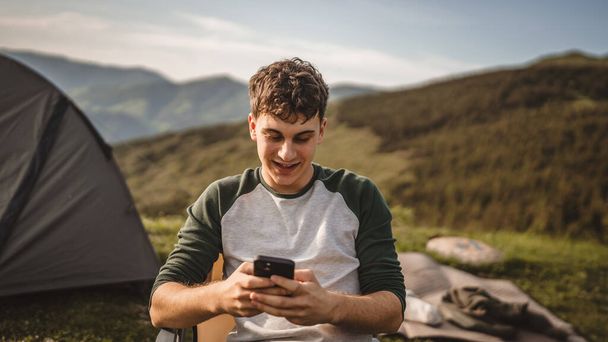 Νεαρό αγόρι κάθεται μπροστά σκηνή χρησιμοποιούν το κινητό τηλέφωνο στο βουνό - Φωτογραφία, εικόνα