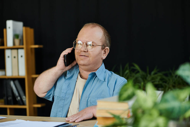 Um homem com inclusividade em óculos senta-se em uma mesa, falando ao telefone. - Foto, Imagem
