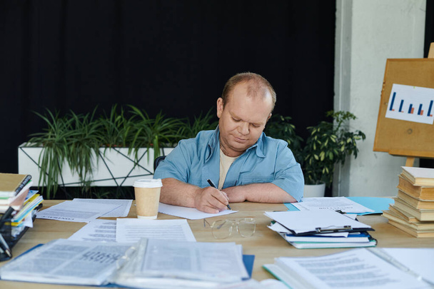 Ένας άντρας με εγκλειστικότητα κάθεται σε ένα γραφείο, γράφοντας σε ένα κομμάτι χαρτί, περιτριγυρισμένος από χαρτιά και ένα φλιτζάνι καφέ.. - Φωτογραφία, εικόνα