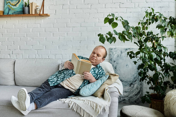 Ein Mann mit Inklusivität entspannt sich auf einer Couch und liest ein Buch. - Foto, Bild