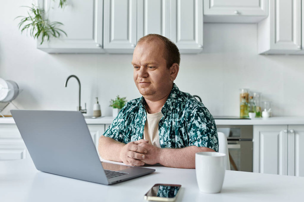 Un hombre con inclusividad se sienta en una mesa en una cocina moderna, trabajando en un ordenador portátil. - Foto, Imagen