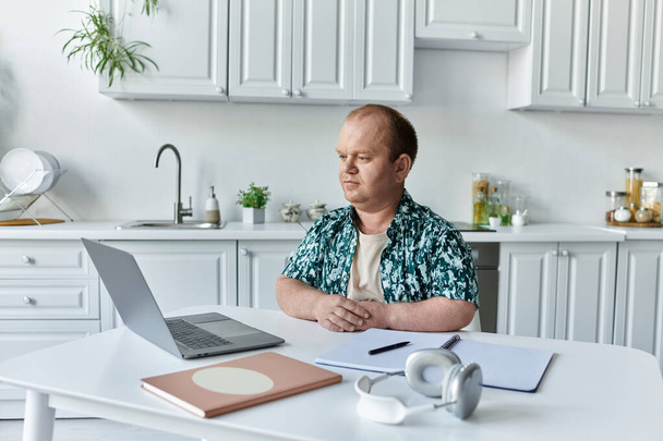Egy inkluzivitású férfi ül egy asztalnál a konyhában, egy laptopon dolgozik.. - Fotó, kép