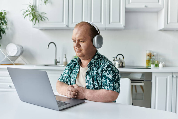 Mężczyzna z inkluzją siedzi przy stole w kuchni, nosi słuchawki i skupia się na laptopie.. - Zdjęcie, obraz