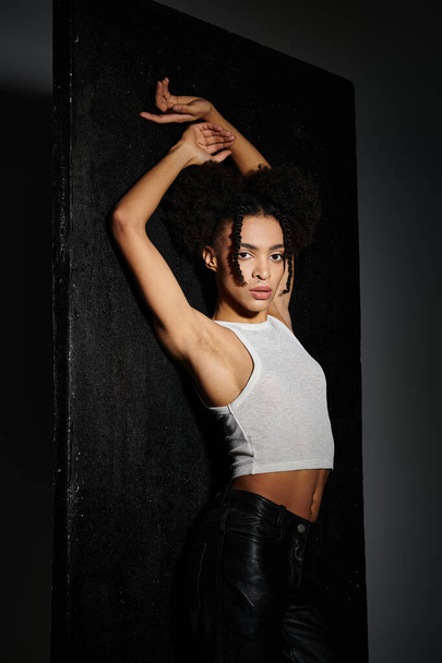 A young African American woman poses against a dark grey backdrop, wearing a white tank top and leather pants. - Photo, Image
