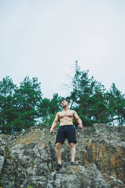 Ein junger Mann mit Bauchwürfeln auf dem Hintergrund des Waldes. Athletischer Mann auf einem Felsen. - Foto, Bild