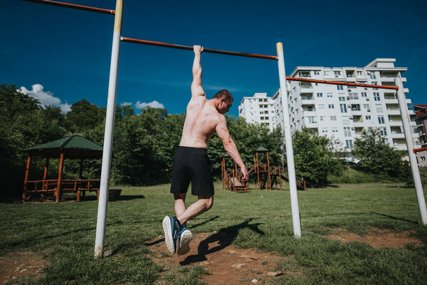 Πλάτη άποψη ενός άνδρα κάνει pull-ups σε ένα αστικό πάρκο σε μια ηλιόλουστη μέρα με πράσινο γρασίδι και δέντρα. - Φωτογραφία, εικόνα