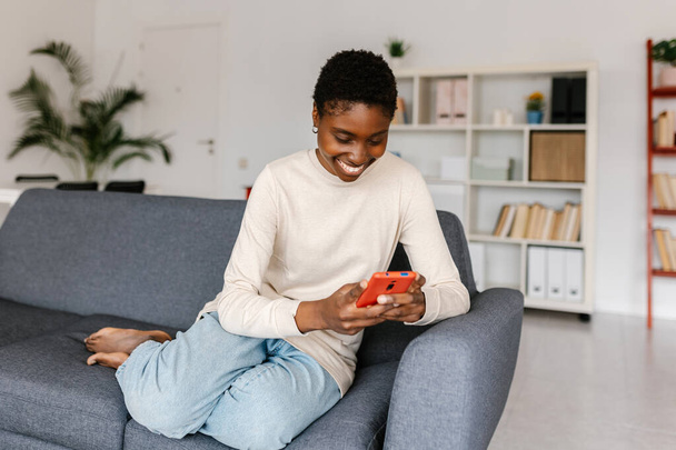 Młoda afrykańska dorosła kobieta uśmiecha się podczas korzystania z telefonu komórkowego, siedząc na kanapie w domu. Koncepcja technologii i sieci społecznych. - Zdjęcie, obraz