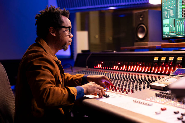 African American ήχου σχεδιαστής χρησιμοποιεί γραφείο ελέγχου με αυτόματη ισοσταθμιστή, μίξερ και faders για να επεξεργαστείτε τη μουσική μετά την παραγωγή. Ηχητικός εμπειρογνώμονας επεξεργασίας ηχογραφήσεις, ανάμειξη κονσόλας. - Φωτογραφία, εικόνα