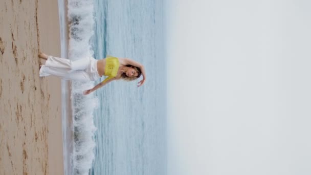 Женщина грациозно движется тело на песчаном пляже вечером вертикально ориентированы. Выразительная курчавая исполнительница, танцующая современную хореографию в передних морских волнах. Танцовщица делает плавные движения на берегу моря - Кадры, видео