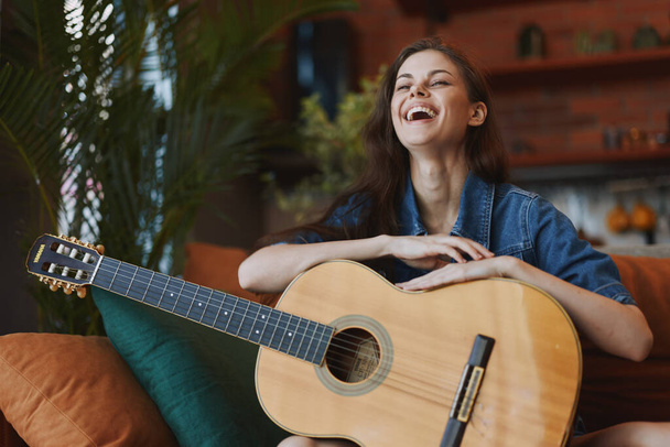 Счастливая женщина сидит на диване с акустической гитарой перед ней, улыбаясь - Фото, изображение