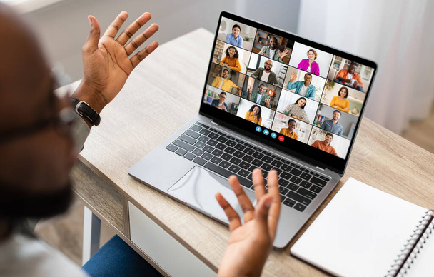 Black man sits at a desk in front of a laptop screen, participating in a video conference meeting. He has both hands raised, gesturing to the other participants in the call. - Photo, Image