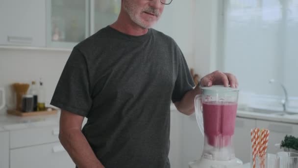 Inclinado retrato de hombre caucásico maduro en gafas haciendo batido saludable de frutas y mirando a la cámara en la cocina - Metraje, vídeo