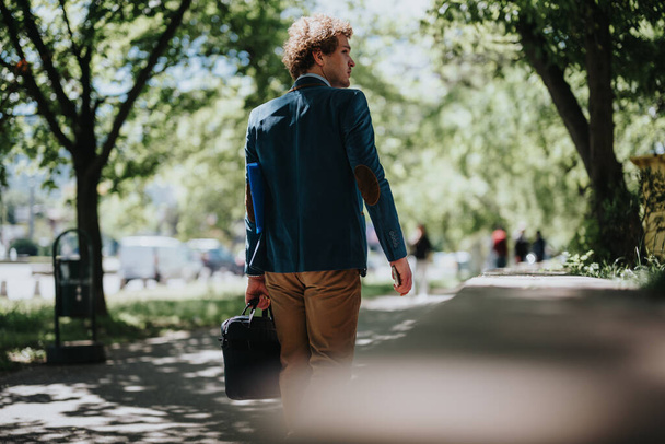 Şık mavi ceketli odaklanmış bir işadamı, elinde siyah bir evrak çantası, profesyonelliği ve kararlılığı simgeleyen, güneşli bir parkta yürüyor.. - Fotoğraf, Görsel