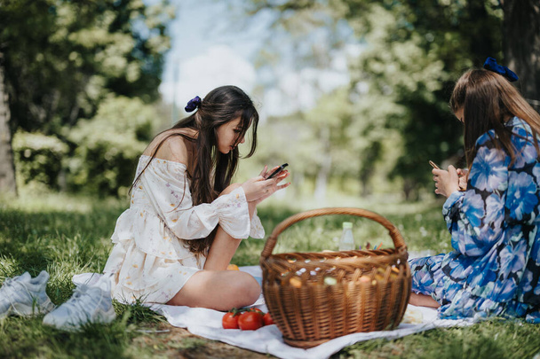 İki genç kadın, muhtemelen kız kardeşler, ağaçlarla çevrili güneşli bir parkta piknik yapıyorlar ve akıllı telefonlar kullanarak meyvelerini paylaşıyorlar.. - Fotoğraf, Görsel