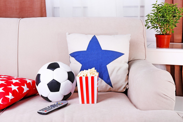 Soccer ball, remote control and box of popcorn on comfortable sofa, indoors - Fotó, kép