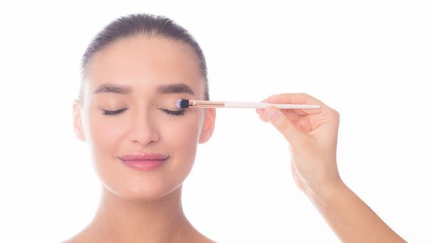 Een vrouw houdt een borstel vast en brengt handig eyeliner aan op haar oogleden. De gefocuste blik op haar gezicht toont precisie en concentratie in het make-up applicatieproces. - Foto, afbeelding