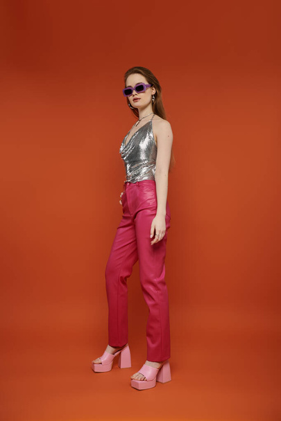 A young woman in a silver top and pink pants poses against a vibrant orange backdrop. - 写真・画像