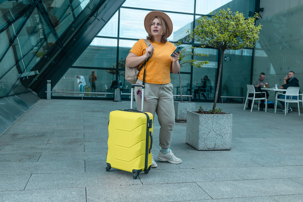 Onnellinen matkustaa Valkoihoinen 30s nainen tyylikäs rento vaatteita ja hattu käyttää mobiili älypuhelin, tarkkailun matkan kohde Internetissä. Nainen odottaa lennon portit kone lennolle, hymyillen on - Valokuva, kuva