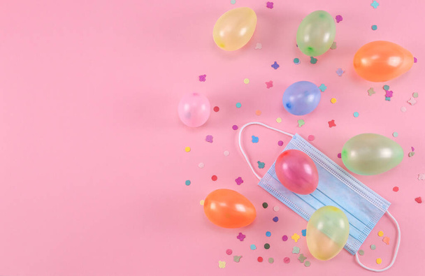 Veel opgeblazen mini ballonnen, een medisch masker en verstrooid papier confetti ligt aan de rechterkant op een roze achtergrond met kopieerruimte in de linker flat lag close-up. - Foto, afbeelding