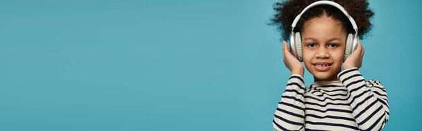 Молодая афроамериканка с вьющимися волосами улыбается, надевая наушники на фоне синего фона. - Фото, изображение