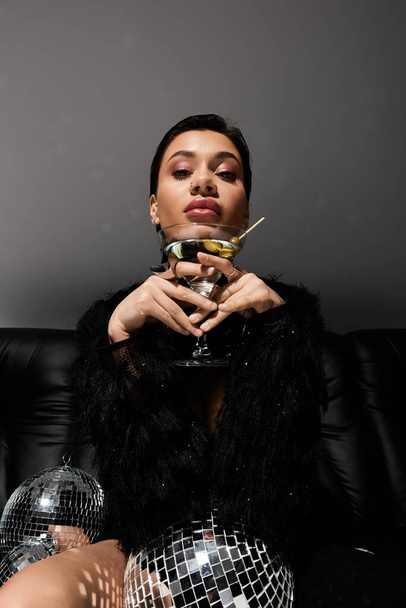 Una mujer en traje sexy sostiene una copa de martini y se sienta en un sofá con una bola de discoteca. - Foto, imagen