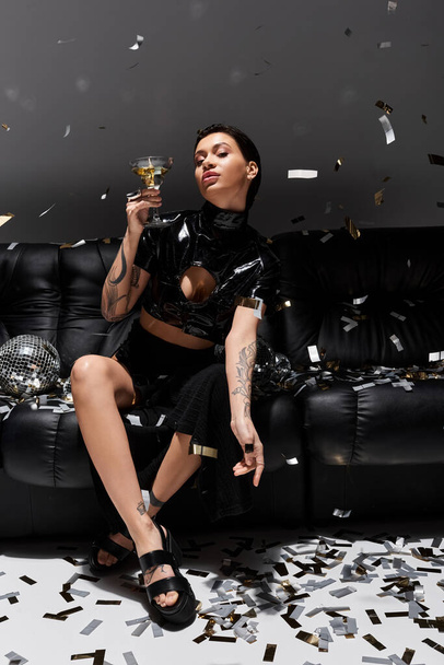 Kobieta w czarnym seksownym stroju, sandały pozuje na kanapie z szampanem, confetti. - Zdjęcie, obraz