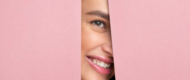 Mujer alegre con su pelo en un moño y con una camisa blanca está asomándose por un agujero en una vibrante pared rosa. - Foto, Imagen