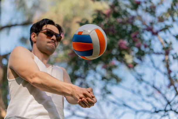 низький постріл на відкритому повітрі в парку з деревами молодий брюнетка хлопець в білій футболці і сонцезахисних окулярах набиває волейбольний м'яч, що прогрівається перед грою дозвілля на вулиці - Фото, зображення