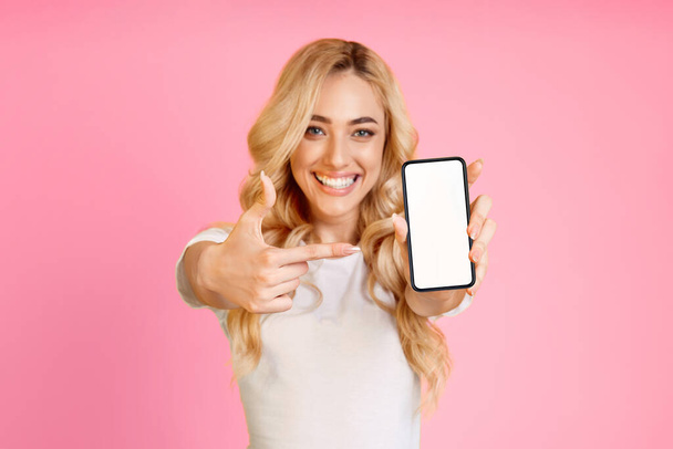 Una giovane donna dai capelli biondi, con una t-shirt bianca, sorride brillantemente alla telecamera mentre sorregge uno smartphone con uno schermo vuoto e lo indica con il dito indice destro. - Foto, immagini