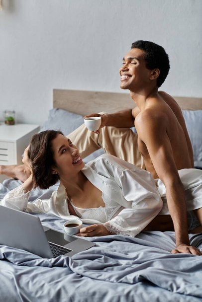 Mladý multikulturní pár si užívá relaxační ráno ve své ložnici, popíjí kávu a sdílí smích. - Fotografie, Obrázek