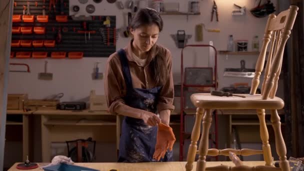 Young woman in an atmospheric workshop preparing to restore an old wooden chair, putting on work gloves. Tools and materials are scattered around as she focuses on her task in the rustic setting. - Footage, Video