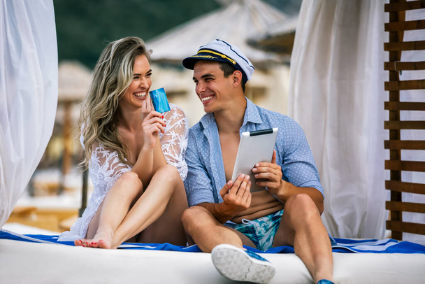 Young beautiful couple ordering online with the card, on the beach. Boyfriend wearing sailor cap. - Photo, Image