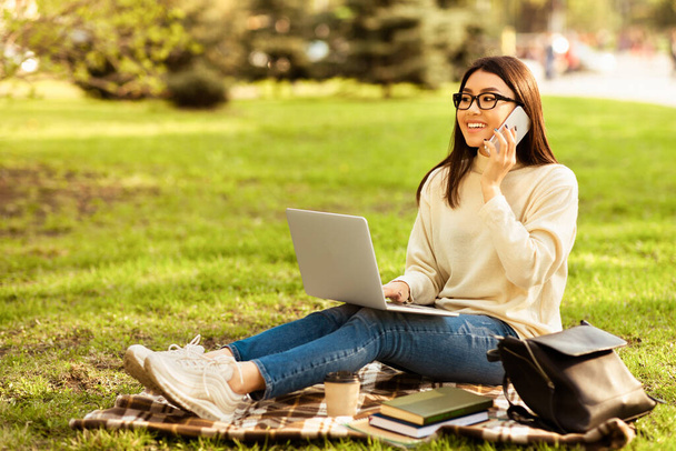 Azjatka siedzi wygodnie na kocu w parku, rozmawiając przez telefon i jednocześnie korzystając z laptopa. Wydaje się zadowolona i produktywna, otoczona książkami. - Zdjęcie, obraz