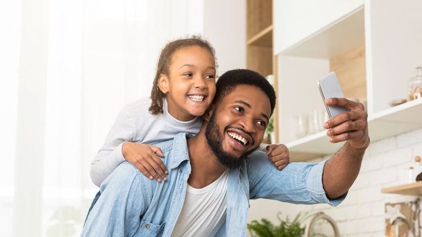 Un père noir souriant prend un selfie avec sa fille dans la cuisine, capturant un moment de joie en famille et de paternité - Photo, image