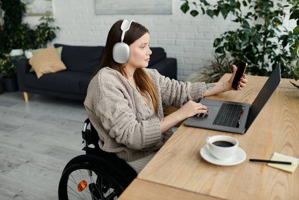Una giovane donna si siede su una sedia a rotelle a una scrivania nella sua casa, lavorando sul suo computer portatile. Indossa le cuffie e guarda il suo telefono.. - Foto, immagini