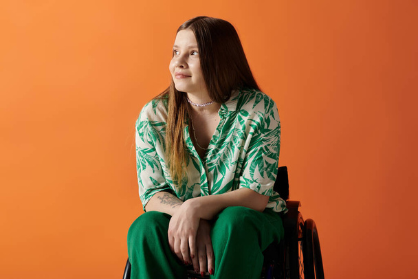 Una joven con el pelo castaño largo y una camisa verde vibrante se sienta en una silla de ruedas sobre un fondo naranja, sonriendo y mirando hacia arriba. - Foto, Imagen