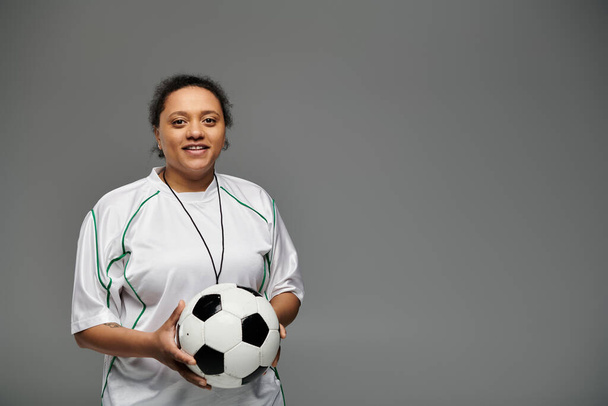 Een atletische vrouw in een witte trui houdt een voetbal vast en glimlacht zelfverzekerd, klaar voor de wedstrijd. - Foto, afbeelding