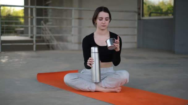 Mujer joven después de la clase de yoga bebe té de un termo en una esterilla de yoga - Metraje, vídeo