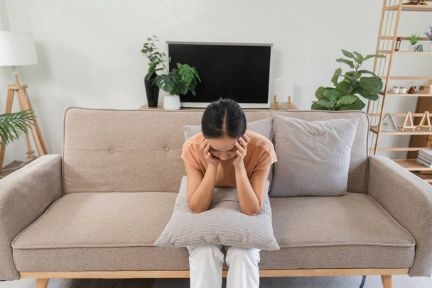 Młoda kobieta siedząca na kanapie, chora na ból głowy, trzymająca poduszkę dla wygody, wyrażająca dyskomfort i ból w domu. - Zdjęcie, obraz