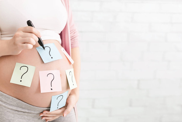 Concepto de elegir el nombre del bebé. Mujer embarazada con signos de interrogación en pegatinas de papel en la barriga, panorama con espacio vacío
 - Foto, imagen