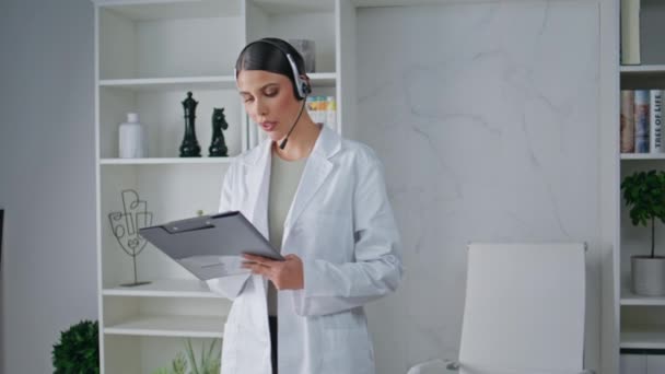 Doctor in headset reading information at clipboard in medicare clinic. Woman therapist in white coat reviewing documentation in polyclinic cabinet. Expert providing attentive service in modern office. - Footage, Video