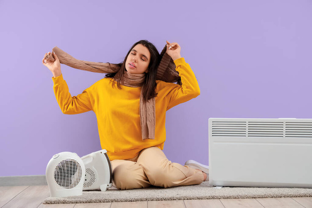 Güzel, terli, kışlık elbiseli, radyatörü ve elektrikli fan ısıtıcısı olan bir kadın mor duvarın yanında oturuyor. - Fotoğraf, Görsel
