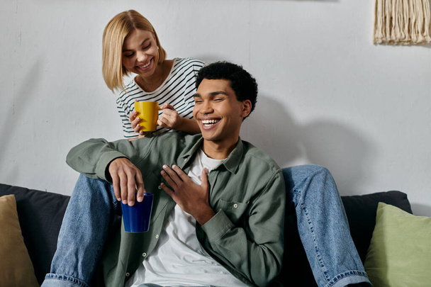 Una giovane coppia interrazziale si rilassa su un divano in un appartamento moderno, godendosi una tazza di caffè e compagnia. - Foto, immagini