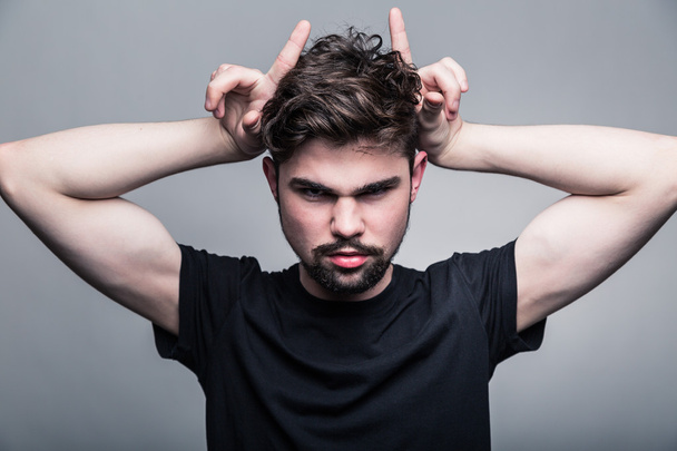 Young man in black T-shirt with fashionable hairstyle shows horns - Photo, Image