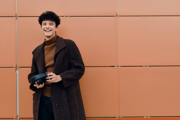 Jovem elegante em casaco marrom e calças pretas em pé na frente da parede laranja com as mãos nos bolsos. estilo urbano na moda. - Foto, Imagem
