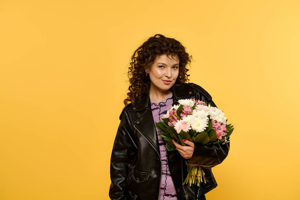 Une jeune femme aux cheveux bouclés portant une veste en cuir noir tient un bouquet de fleurs roses et blanches sur fond jaune. - Photo, image