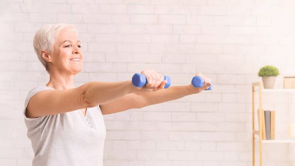 高齢の女性は積極的に白い部屋で運動しています, 彼女の筋肉を強化し,彼女の体力を向上させるためにダンベルを持ち上げます - 写真・画像