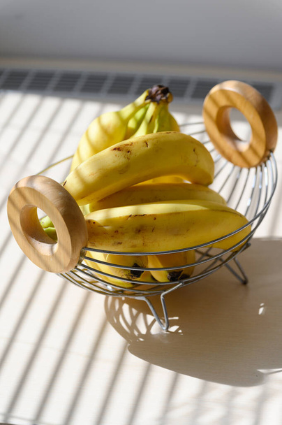 Αρκετές ώριμες μπανάνες σε καλάθι με φρούτα. Θέα από ψηλά. Σκιά από τις περσίδες στο φρούτο. - Φωτογραφία, εικόνα